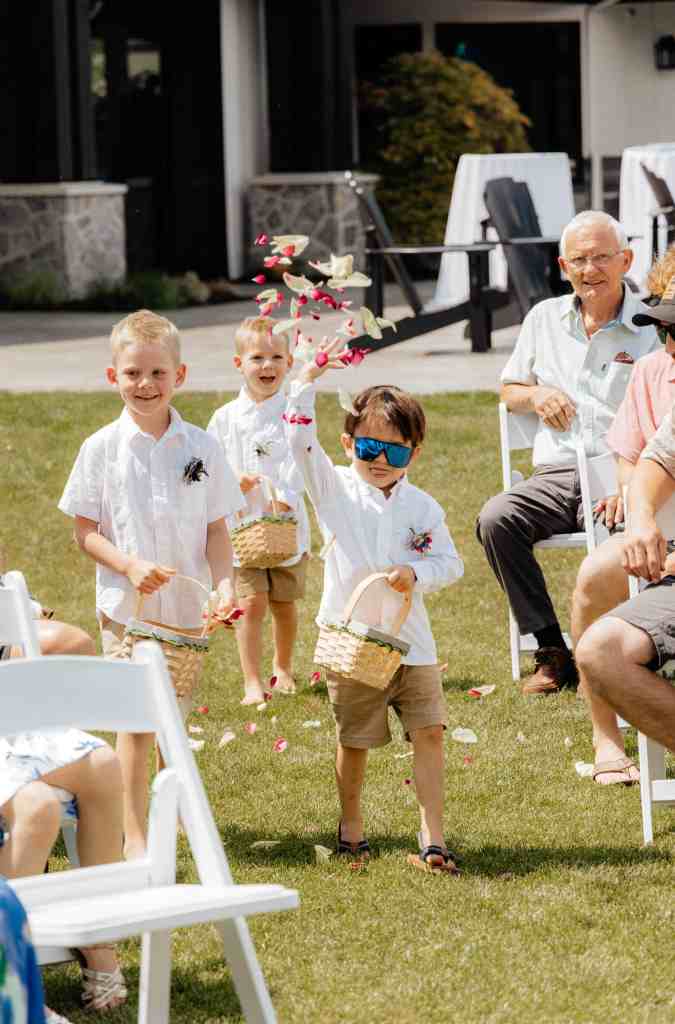 A funny kid in sunglasses throws flower petals before the bride walks down the aisle at a backyard wedding in Kelowna, BC