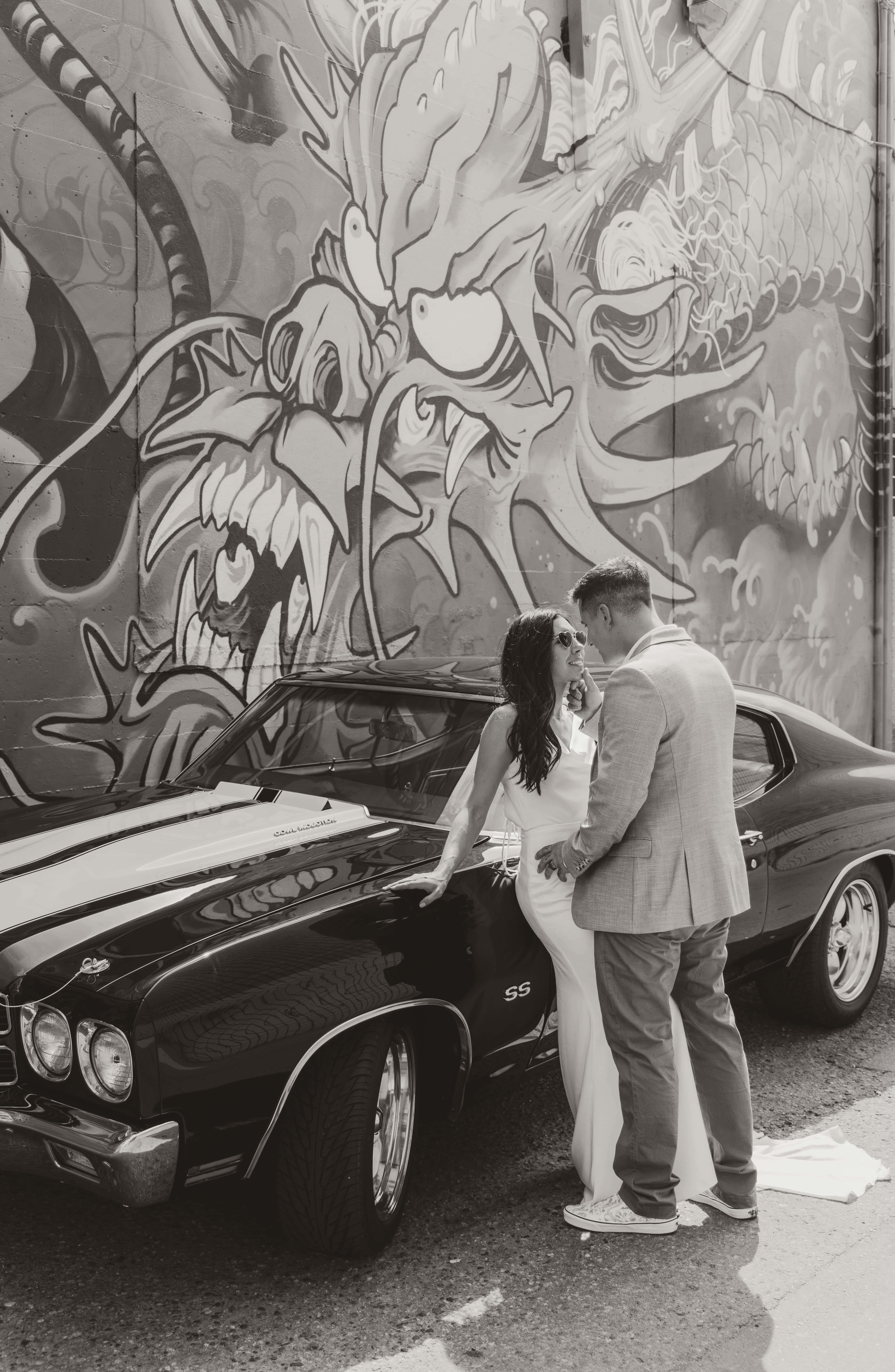 A black and white urban photo of a wedding couple with their vintage car in front of a mural in downtown Kelowna, BC, Okanagan
