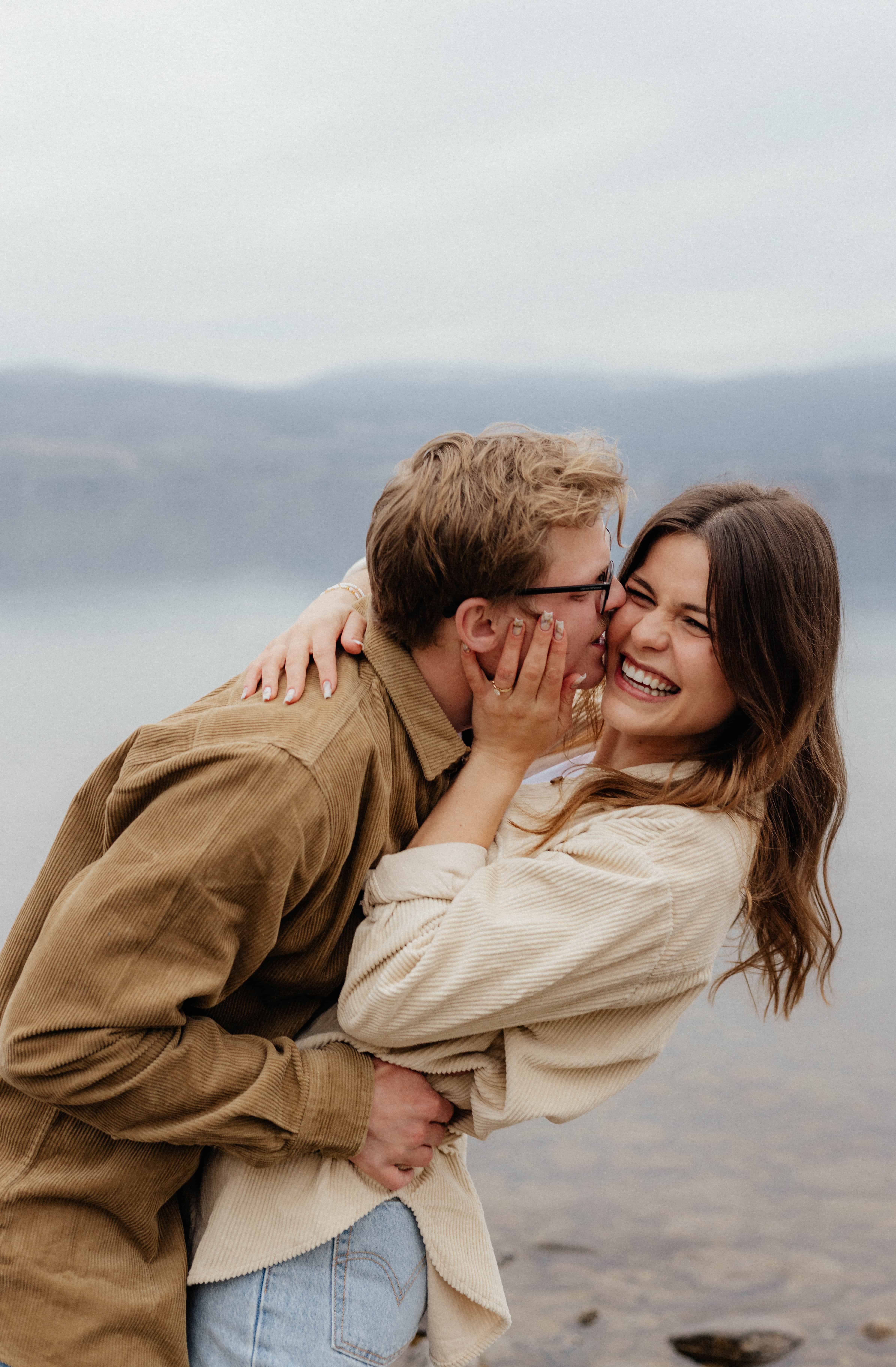 A man playfully kisses his fiancée on the cheek while she laughs during their engagement session at Cedar Creek Park, Kelowna