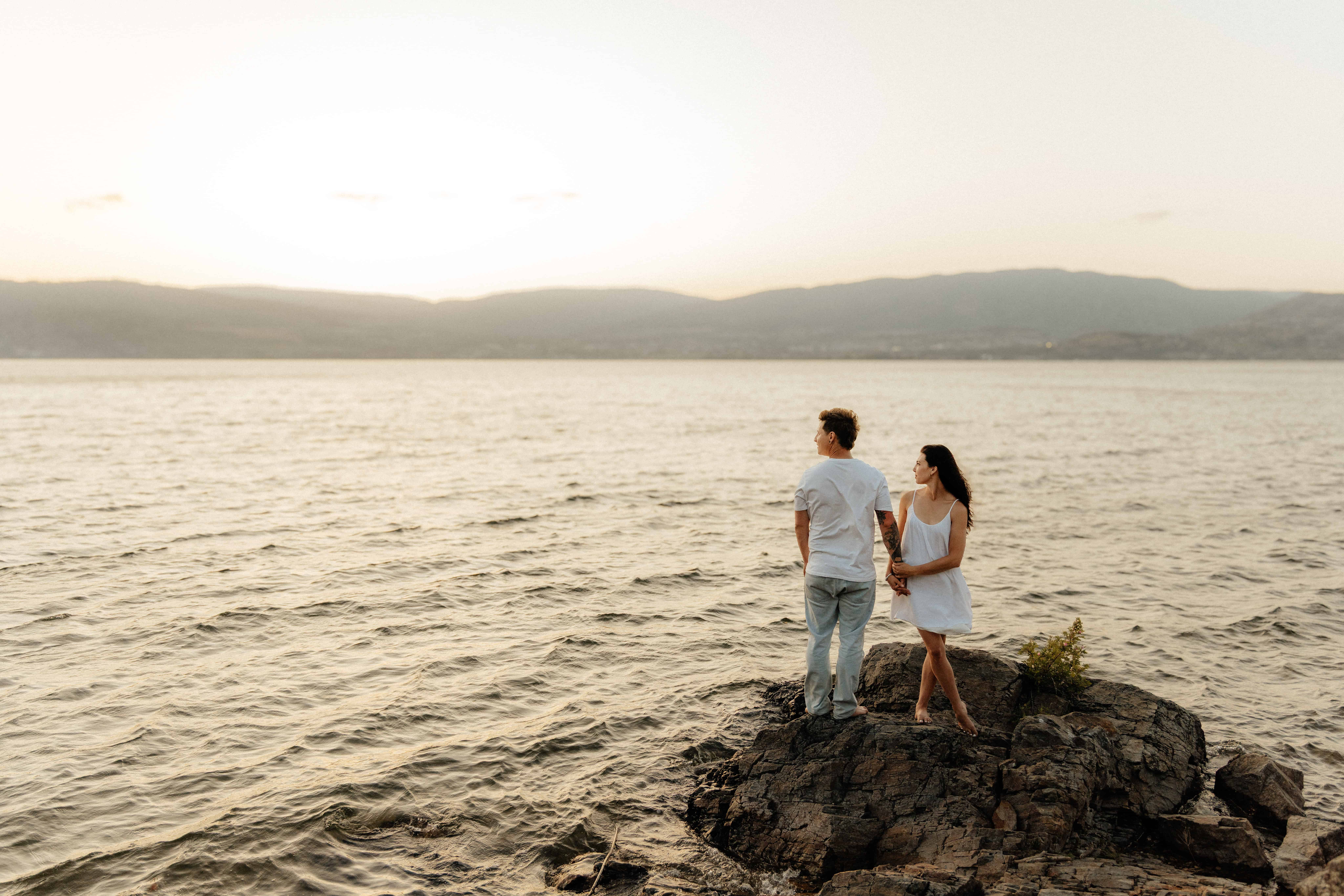 A beautiful couple stands on a rock admiring the view of the Okanagan lake at Bertram Creek Park in Kelowna, BC