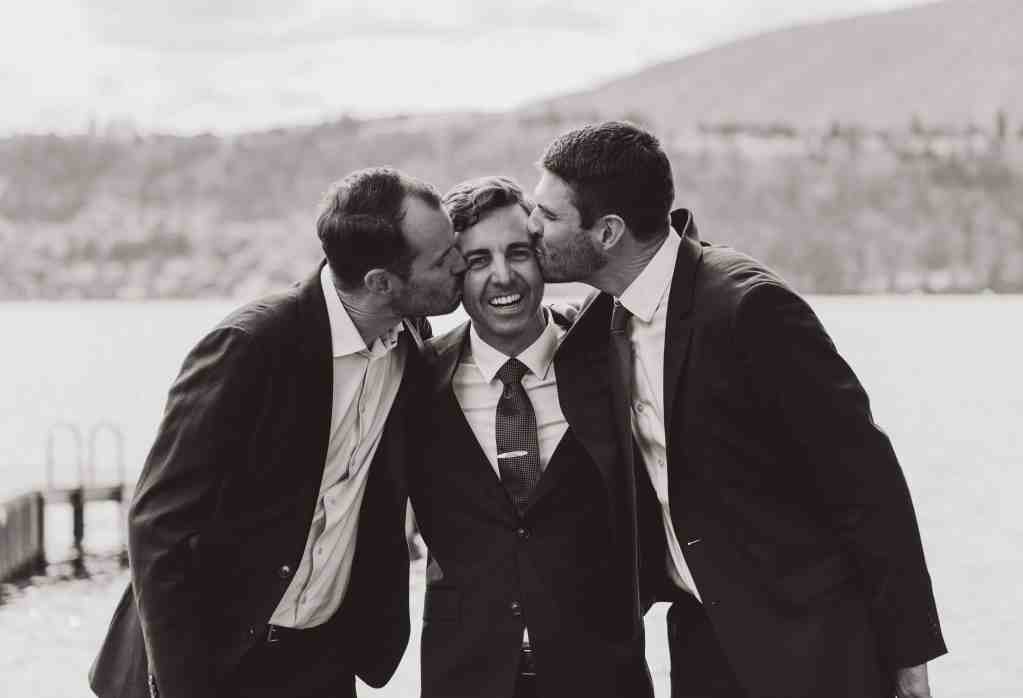 A groom laughs while his two brothers kiss him on either cheek in a classic black and white wedding photo in Kelowna, BC