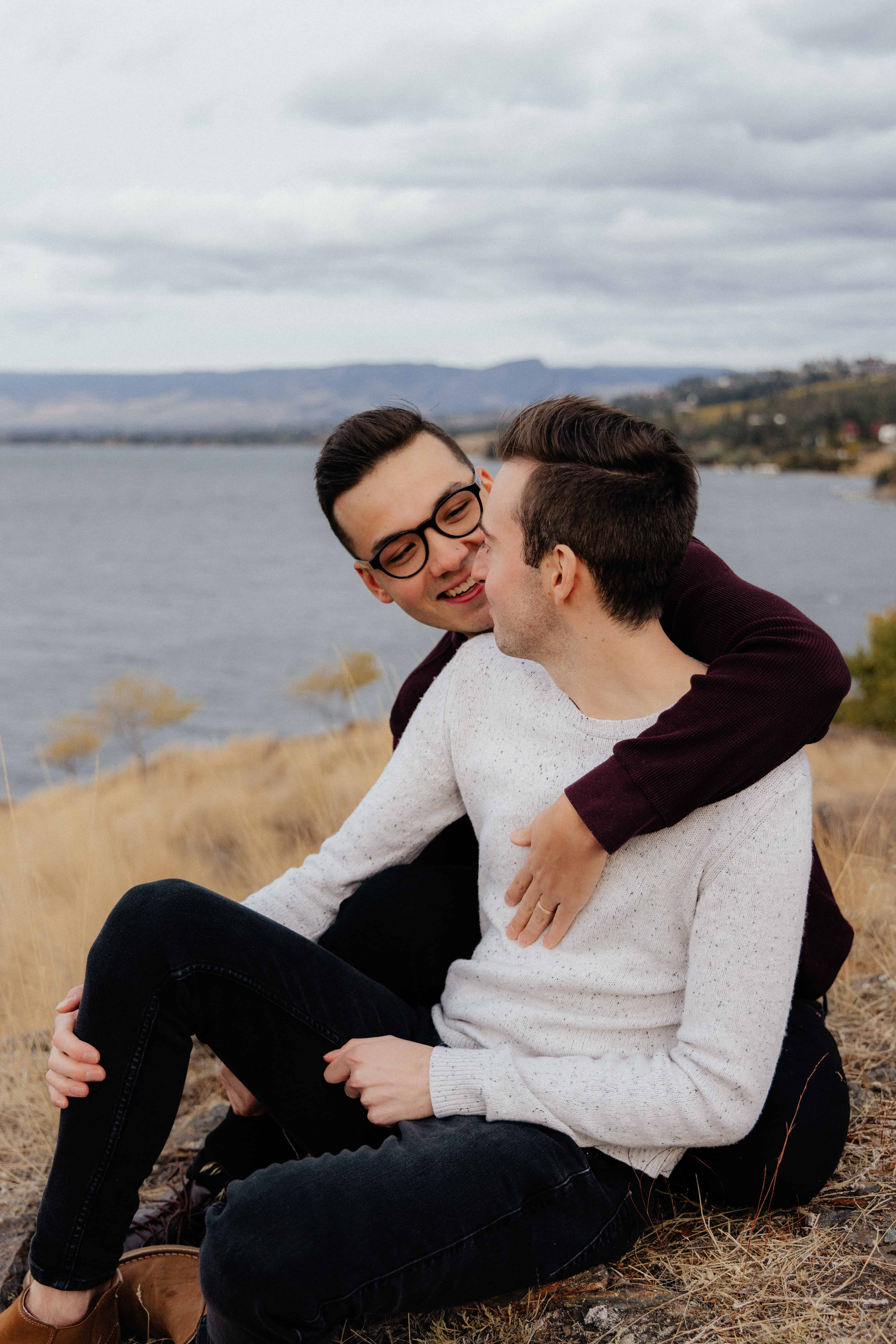 A same-sex couple hug and laugh during their stunning engagement photoshoot at Bertram Creek Park overlooking Lake Okanagan in Kelowna