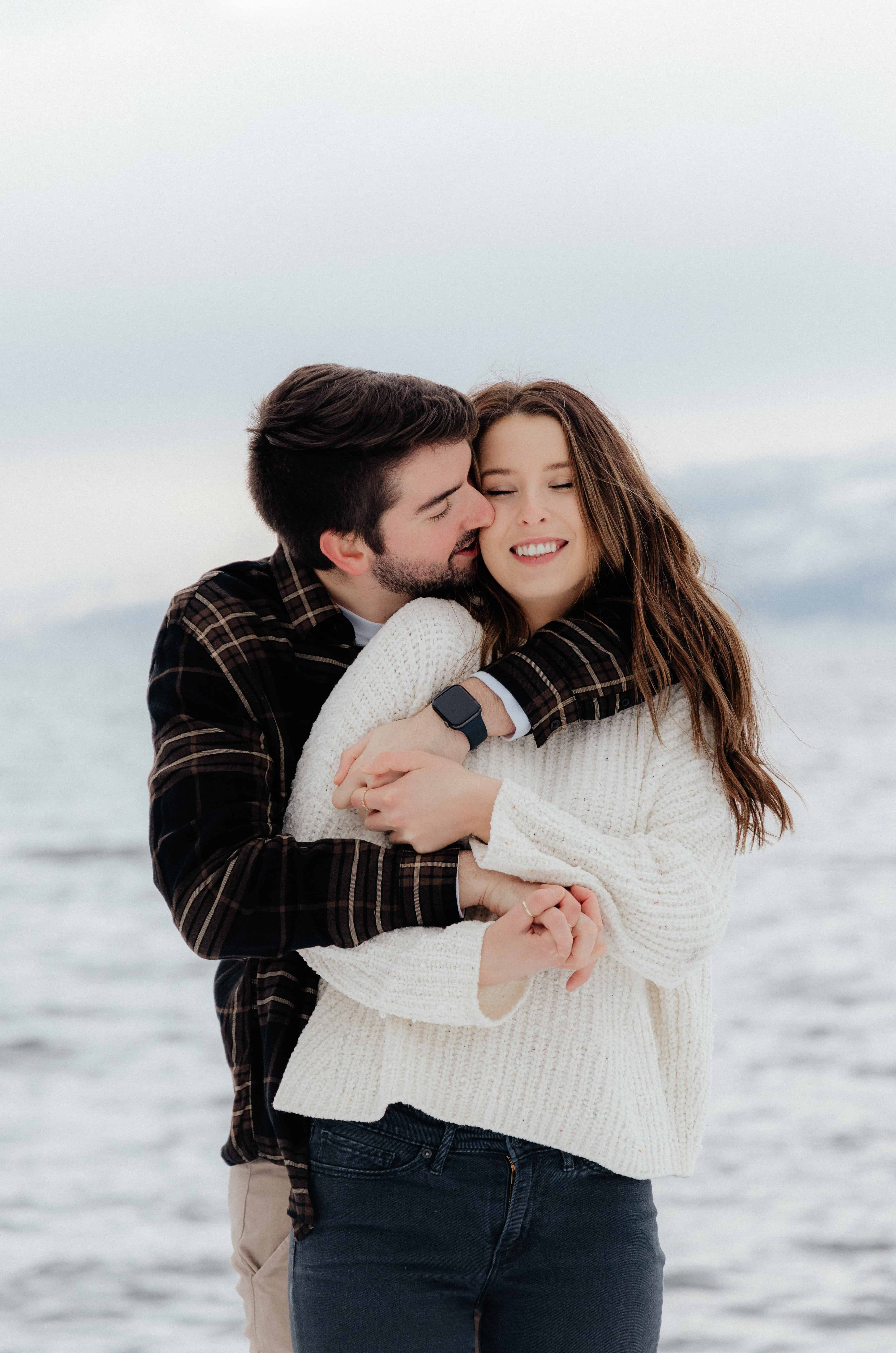 A man embraces his fiancée as she laughs during their winter engagement session in Peachland, BC