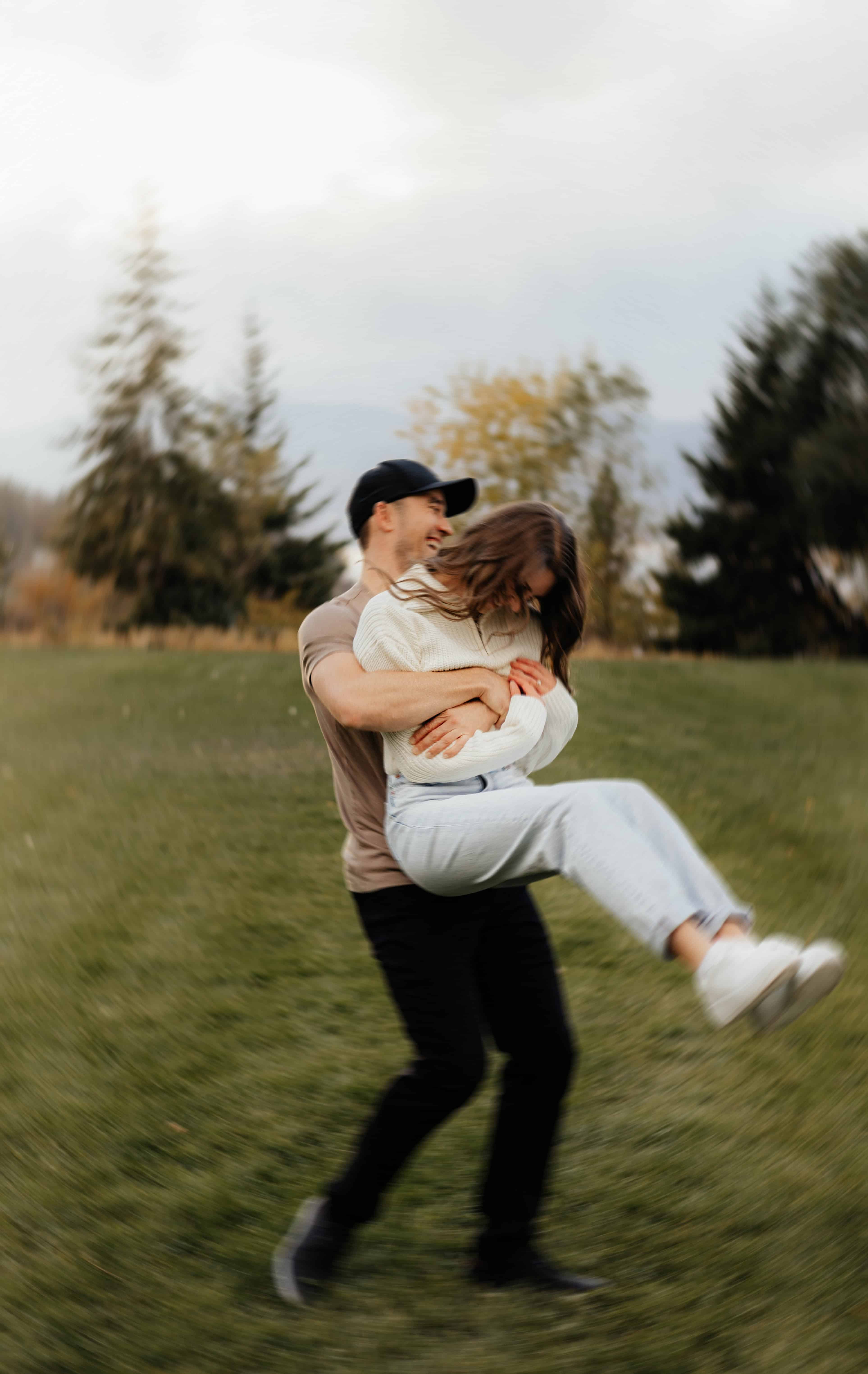 A man lifts and twirls his fiancé in a green field at Bertram Creek Park in Kelowna, Okanagan