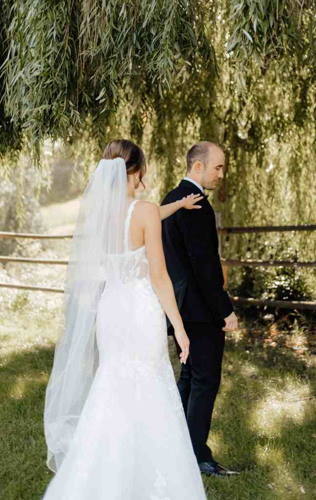 A bride taps her groom on the shoulder for a first look under a willow tree at Summerhill Winery, Okanagan