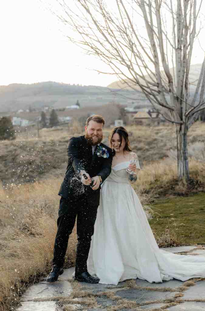 A groom pops a bottle of champagne while his bride giggles as the sun sets behind them at Sanctuary Gardens, West Kelowna. A classic wedding photography shot for beginners.
