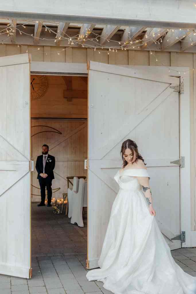 A bride adjusts her gown before walking down the aisle to greet her groom as he can be seen in the background at Sanctuary Gardens in West Kelowna