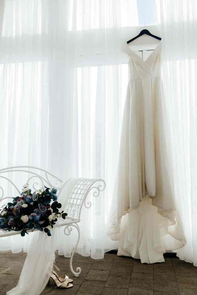 A beautiful photo of a bride's wedding gown hanging in front of a window with white curtains beside her gorgeous bouquet at Sanctuary Gardens, West Kelowna