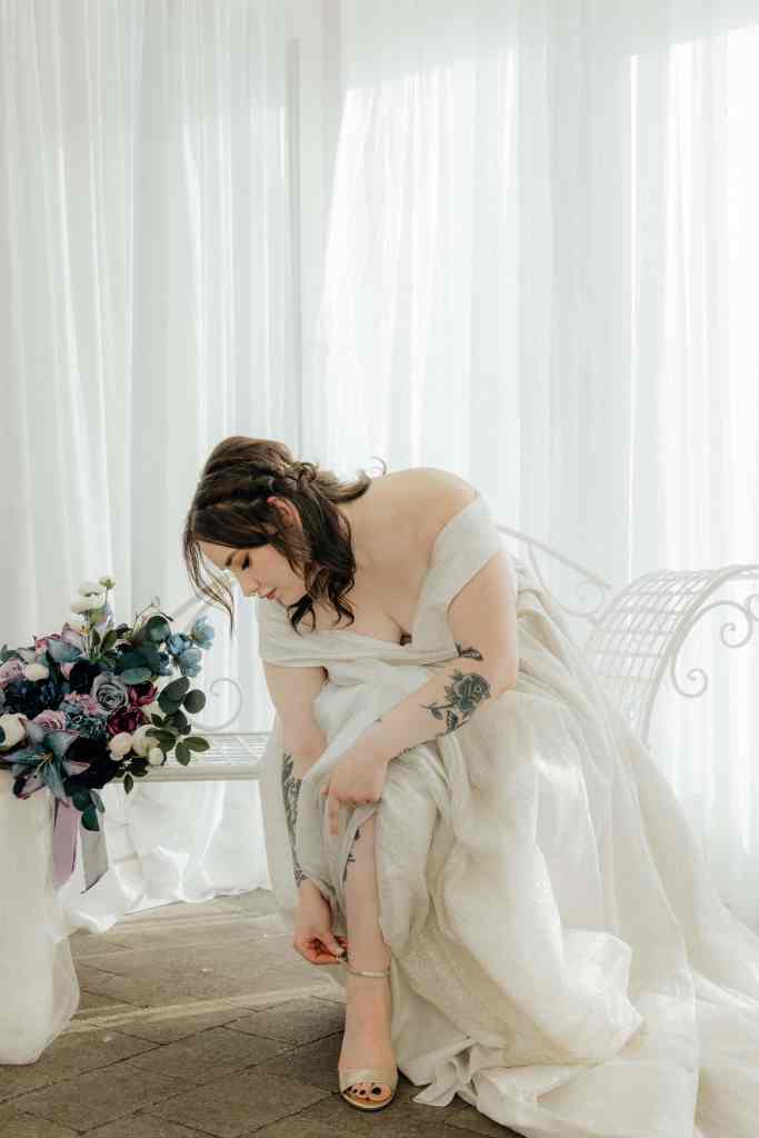A bride leans over to buckle her shoe in a bright white room in Sanctuary Gardens, West Kelowna