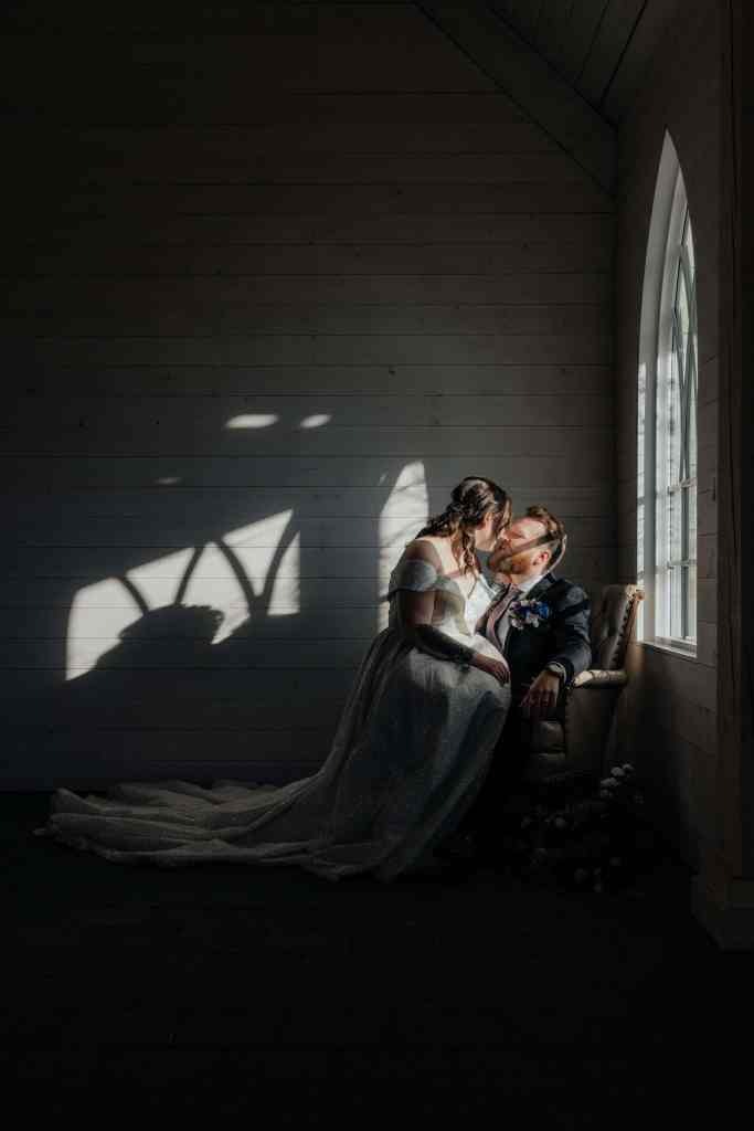 A bride sits on her groom's lap lit by the light of a single window as they share a kiss during their elopement at Sanctuary Gardens in the Okanagan