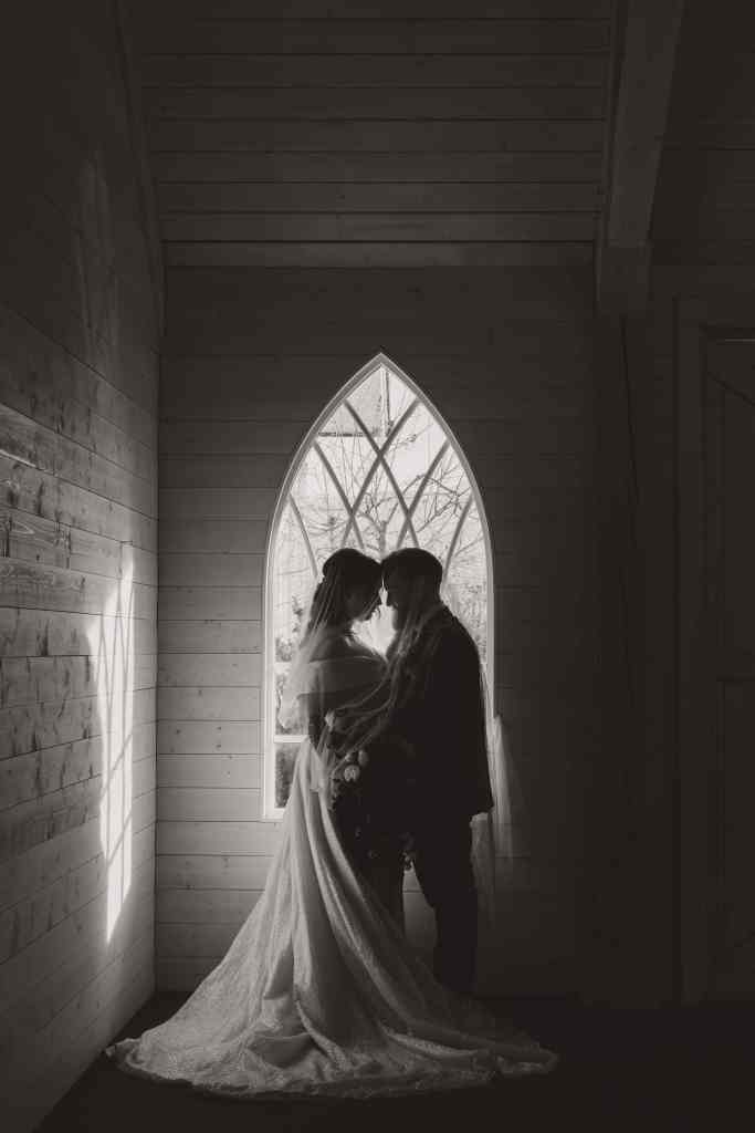 A stunning moody wedding portrait of a bride and groom draped in a veil in front of an ornate window at Sanctuary Gardens