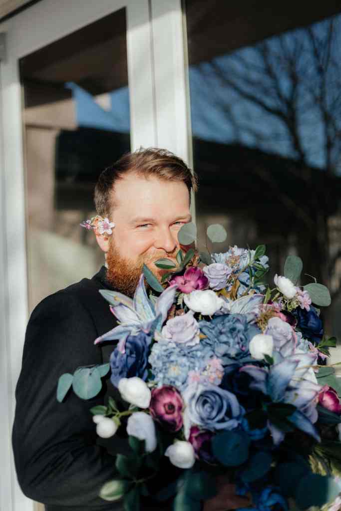 A cute in-the-moment photograph of a groom holding his bride's bouquet with a flower behind his ear at Sanctuary Gardens