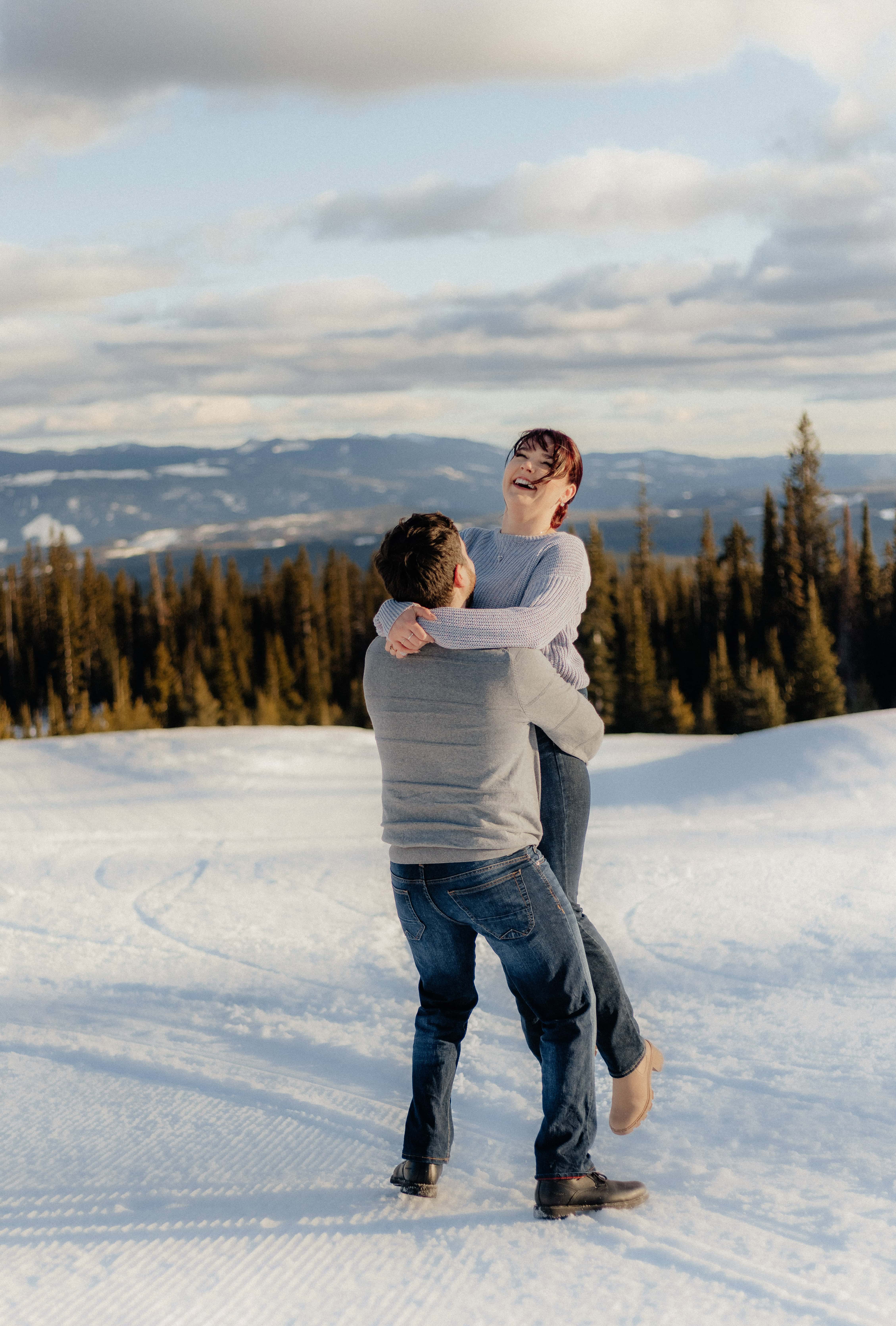A man playfully picks up and spins his fiancée during their snowy engagement photoshoot overlooking the Okanagan mountains at Big White Ski Resort