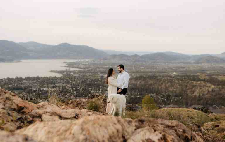 A couple and their dog pose for a photo overlooking Kelowna, BC during their engagement session