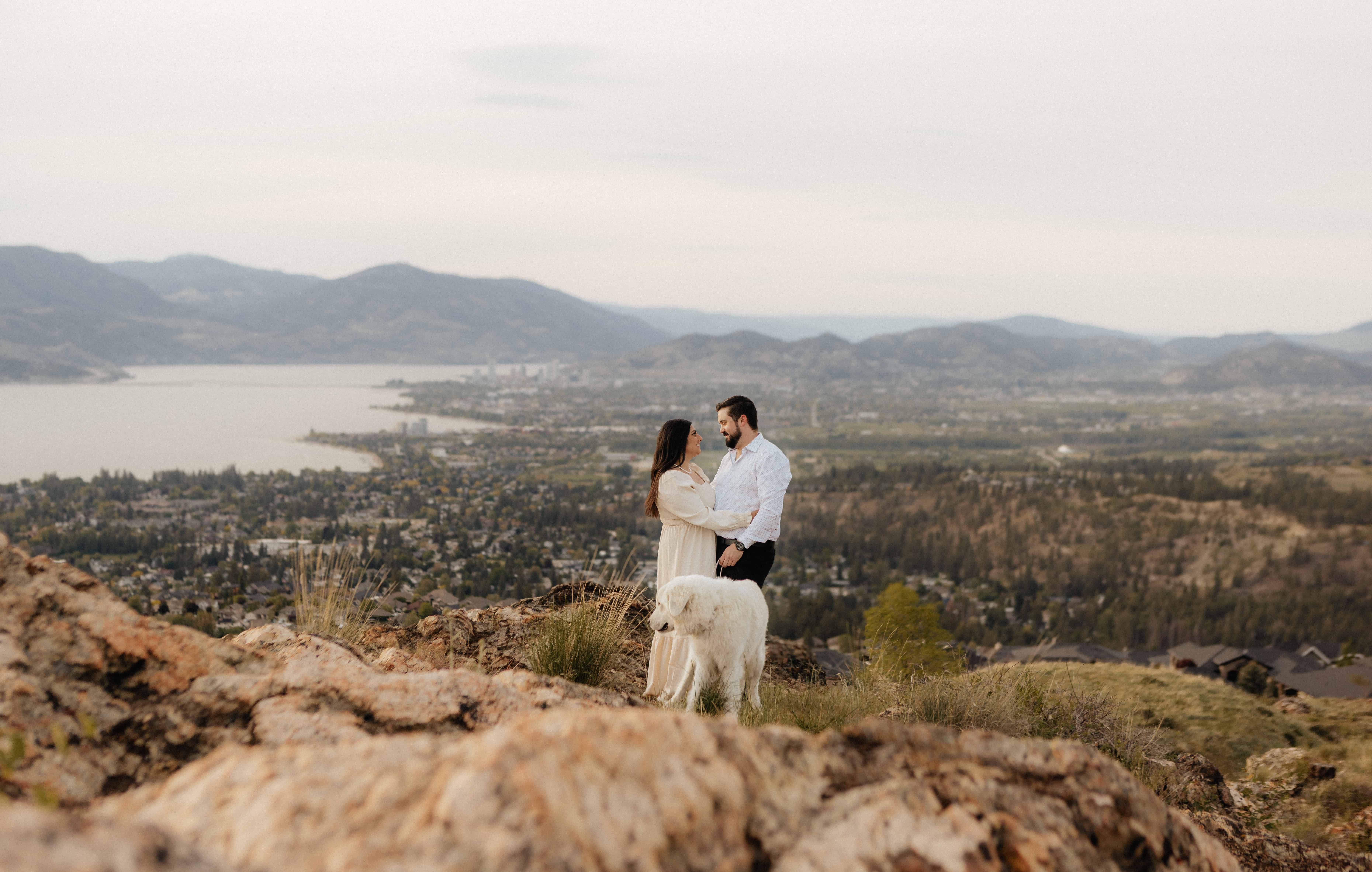 A couple and their dog pose for a photo overlooking Kelowna, BC during their engagement session