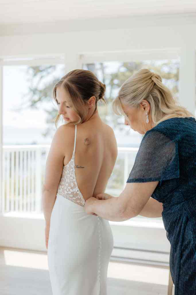 A mother buttons up her daughter's wedding dress before the couple exchanges vows at the Cove at Otter Point for a beautiful Vancouver Island Wedding